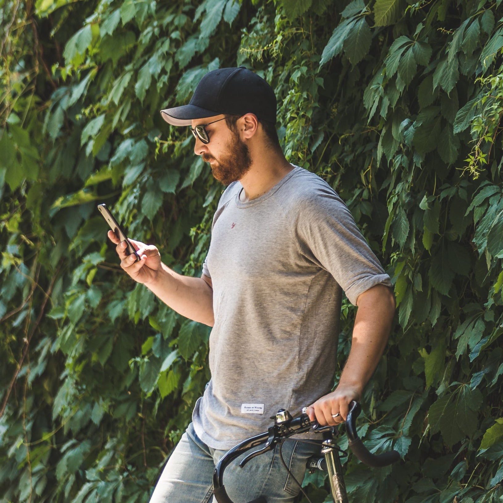 Man with a bicycle using a phone against a green leafy wall for testimonial of slowing down aging and feeling younger with vitamins, with regrowing hair with topical solution and oral prescription tablet pills dutasteride and minoxidil.