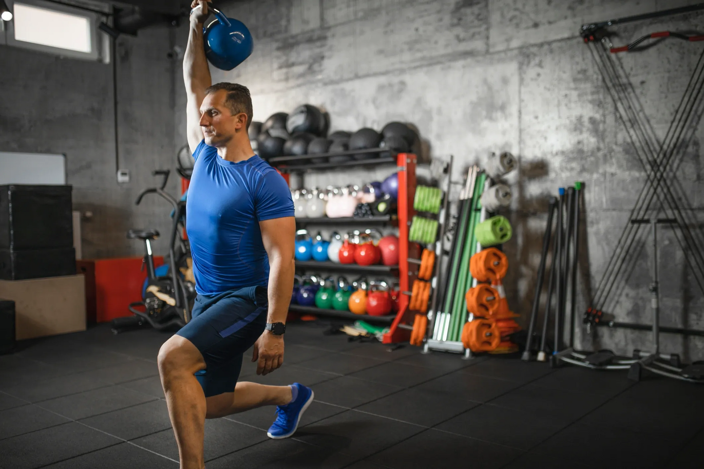 Man lifting a blue kettlebell in a gym setting with various equipment in the background.